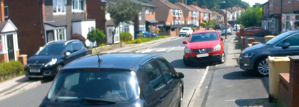 A residential street with cars partially blocking the footways on both sides of the road. A car in the foreground is parked correctly on the carriageway.