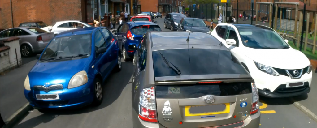 A narrow residential street with a local primary school on the right hand side. The street is full of cars being driven and parked, with some people driving their cars on the footway outside the school gates to squeeze past others.